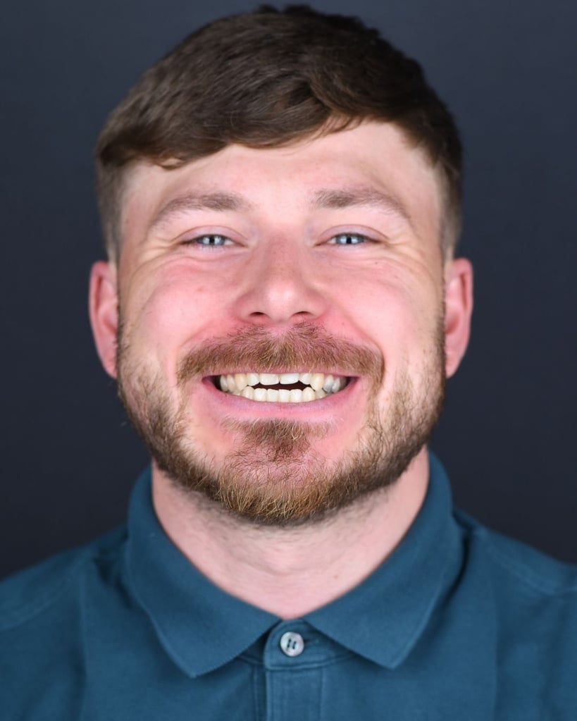 Portrait of a young man with short blond hair and a trimmed beard smiling naturally, showing slightly spaced natural teeth, wearing a teal collared shirt at Smile Center Turkey.