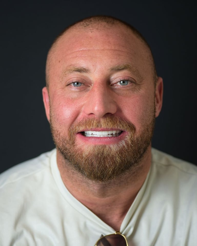 Portrait of a bald man with a trimmed beard and moustache, wearing a white T-shirt and sunglasses around his neck, smiling warmly and showing bright white teeth after dental restoration at Smile Center Turkey.