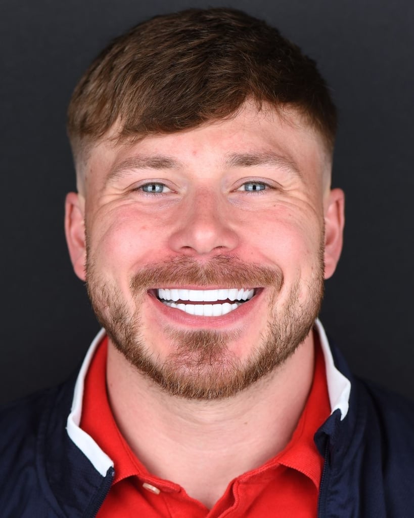 Portrait of a young man with short blond hair and a trimmed beard smiling broadly to show bright white teeth, wearing a red polo shirt and navy jacket at Smile Center Turkey.