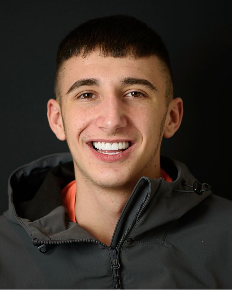 Portrait of a young man with dark brown hair wearing a grey jacket over an orange shirt, smiling broadly and showing bright white teeth after dental restoration at Smile Center Turkey.