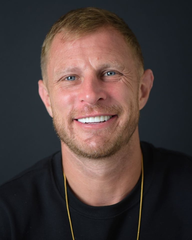 Portrait of a man with short light brown hair and a trimmed beard, wearing a black shirt with a yellow strap, smiling widely and showing straight white teeth after dental restoration at Smile Center Turkey.