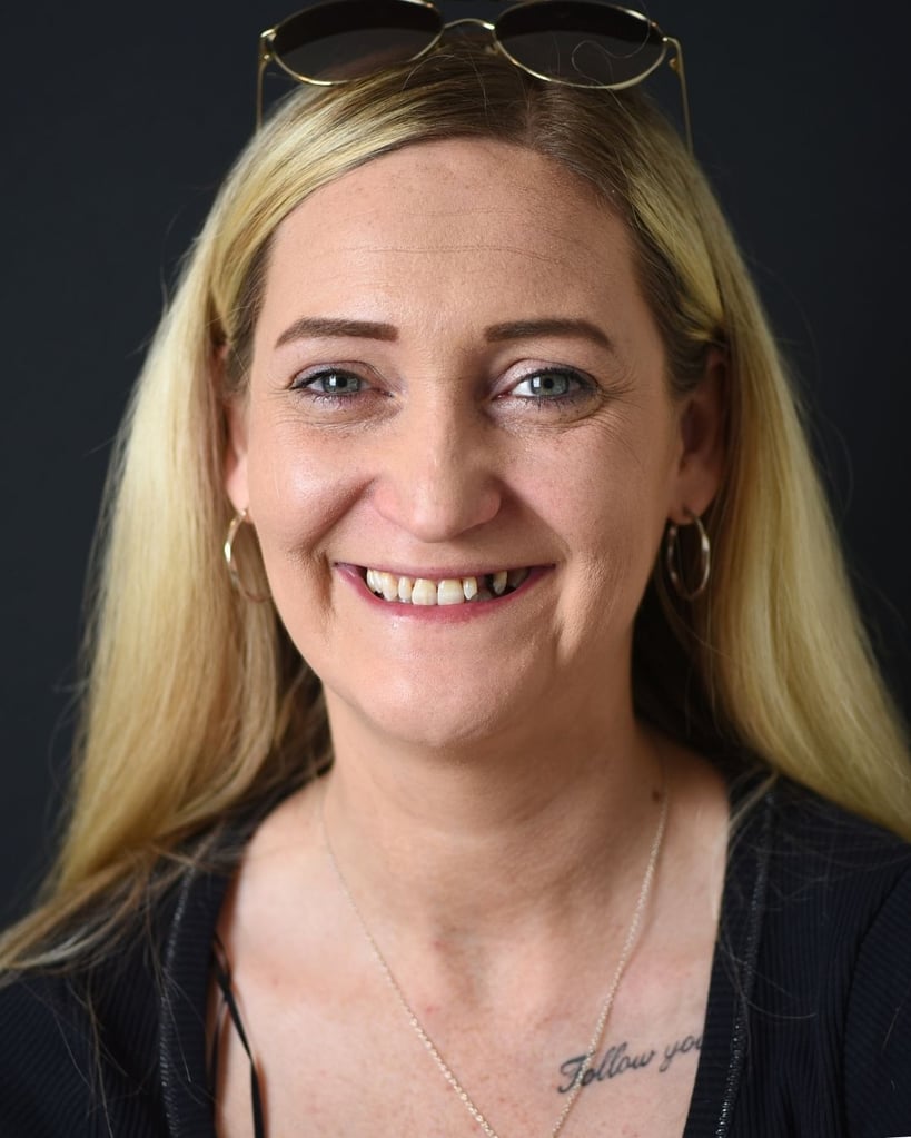 Portrait of a woman with long blonde hair wearing glasses atop her head and a black blazer, smiling slightly and showing missing and discoloured front teeth before dental treatment at Smile Center Turkey.