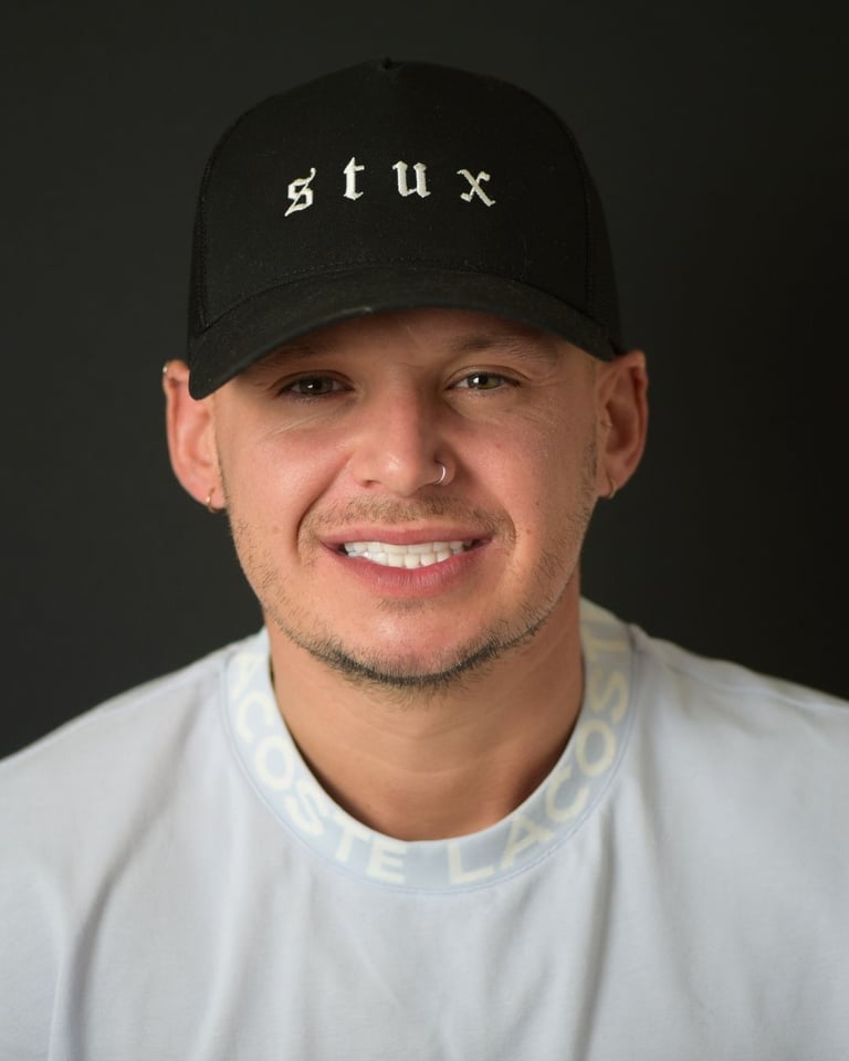 Portrait of a man wearing a black cap with 'stux' lettering, a white Lacoste T-shirt, and a nose ring, smiling widely and showing bright white teeth after dental restoration at Smile Center Turkey.
