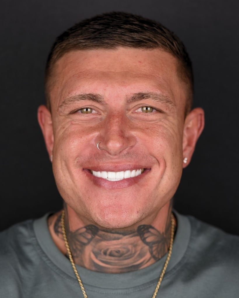 Portrait of a young man with a nose ring and tattooed neck, short hair, and bright white smile against a dark background at Smile Center Turkey.