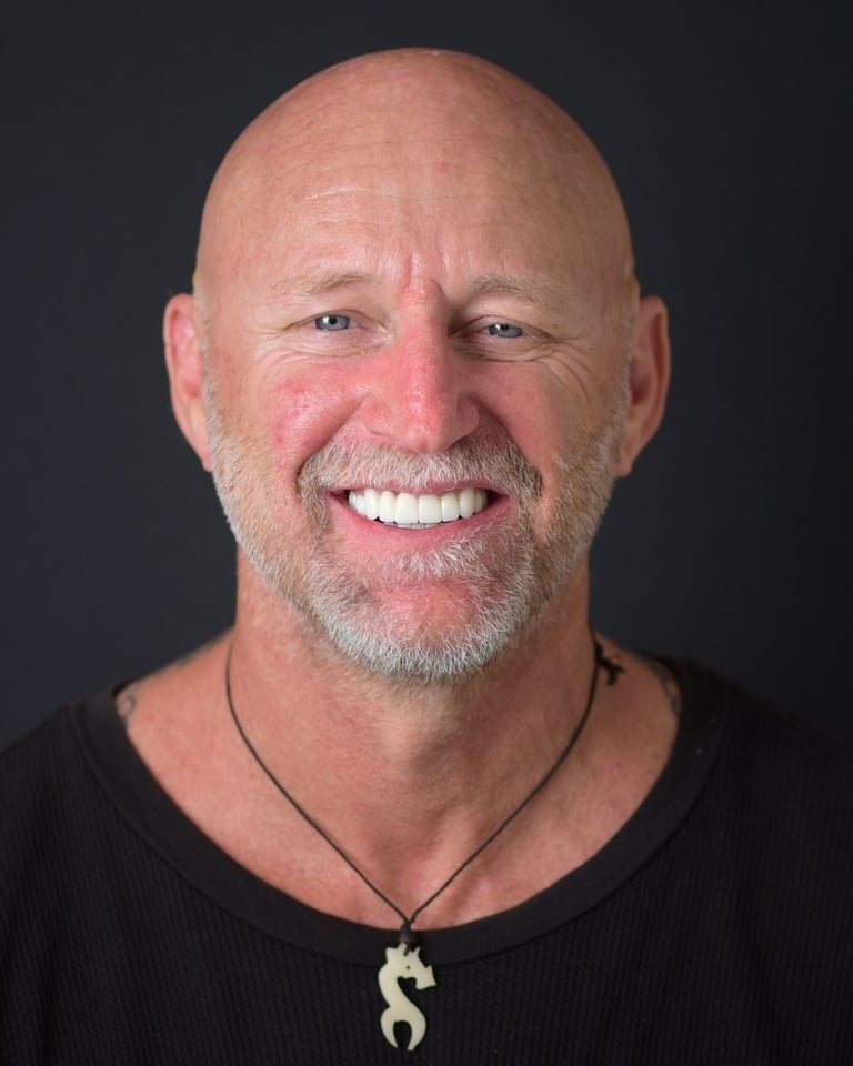Portrait of a bald man with a trimmed grey beard, wearing a black top and a pendant necklace, smiling widely and showing bright white teeth after dental restoration at Smile Center Turkey.