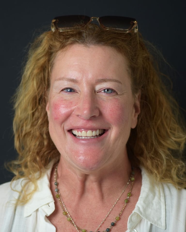 Portrait of a woman with curly blonde hair and glasses on her head, wearing a white shirt and multicolored beaded necklace, smiling widely and showing bright white teeth after dental restoration at Smile Center Turkey.